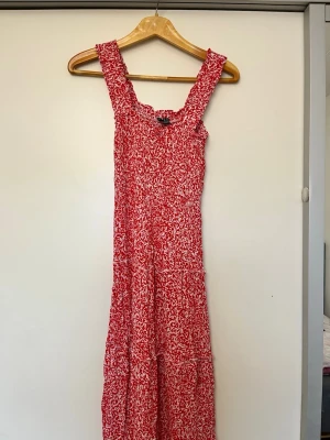 Röd och vit långklänning med mönster - Långklänning i rött och vitt med blommigt mönster och breda axelband. Klänningen har smock upptill och faller snyggt längs kroppen. Perfekt för varma dagar och sommarens alla tillfällen.