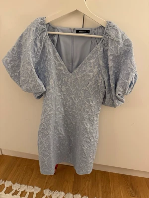 Ljusblå kort klänning från Gina Tricot - Supersöt ljusblå kort klänning från Gina Tricot med puffärmar och v-ringning. Klänningen har ett vackert broderat blommönster och struktur i tyget som ger en exklusiv känsla. Perfekt för sommarens alla tillfällen! Endast använd i ett par timmar. 