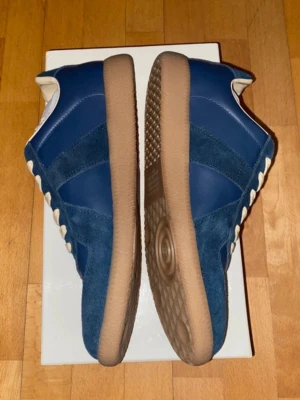 Blå sneakers från Maison Margiela - Snygga blå sneakers från Maison Margiela med ovandel i mocka och läder, vita snören och klassisk gummisula i brunt. Skorna har en låg silhuett och retroinspirerad design som passar perfekt till streetwear och casual outfits.