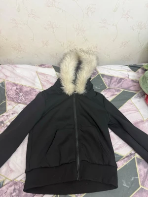 Svart hoodie med fuskpäls på huvan - Snygg svart hoodie med dragkedja och stor huva kantad med mjuk fuskpäls. Två fickor framtill och ribbade muddar vid ärmslut och nederkant. Perfekt för dig som vill ha en chill och trendig look.