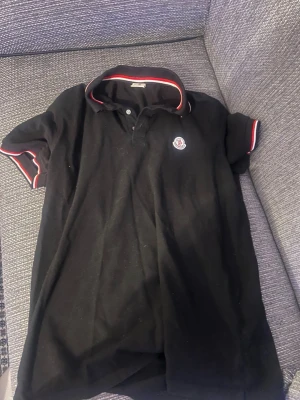 Svart Moncler piké med röd/vita detaljer - Säljer en svart pikétröja från Moncler med korta ärmar och klassisk krage. Tröjan har rödvita detaljer på krage och ärmslut samt Moncler-logga på bröstet. Perfekt till en clean och sportig look.