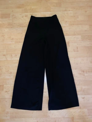 Vida svarta byxor från Forever 21 - Säljer ett par svarta byxor från Forever 21 med hög midja och vida ben. Byxorna har en minimalistisk design utan synliga fickor eller detaljer och är tillverkade i ett mjukt tyg som ger en snygg siluett.