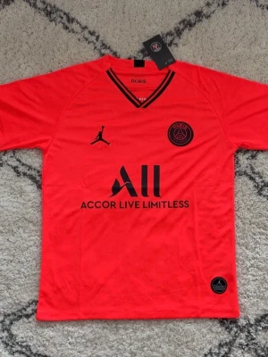 Röd PSG matchtröja Jordan x Nike - Snygg röd PSG matchtröja med svarta detaljer och loggor från Jordan och Paris Saint-Germain. Tröjan har V-ringning med svarta ränder, korta ärmar och är tillverkad i ett lätt och ventilerande material. Perfekt för fotboll eller träning.