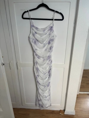 Lång vit-lila klänning - Säljer en lång klänning med tunna axelband i vitt med lila mönster. Klänningen har draperad effekt och omlottdetaljer längs hela längden. Perfekt för sommarens alla tillfällen och har en feminin, elegant look.