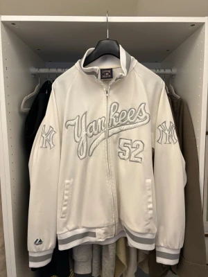 Vit Yankees jacka - Oldschool jacka från Cooperstown Collection med Yankees brodyr på bröstet och ärmarna samt siffran 52. Jackan har ribbade muddar och krage i vitt och grått, dragkedja framtill och två fickor. Perfekt för dig som gillar sportig stil och baseball.