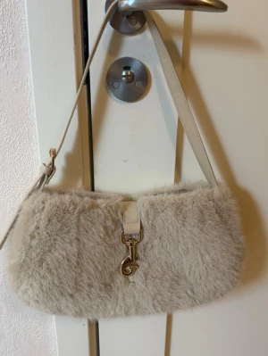 Beige fluffig axelremsväska - Supersöt beige handväska med fluffig pälsimitation och justerbar axelrem. Väskan har ett dekorativt spänne i metall framtill och en mjuk, rundad form. Perfekt accessoar för att lyfta din outfit.