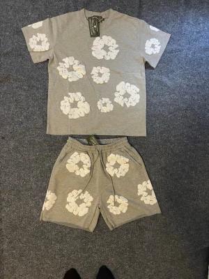 Grått Denim tear set - Snyggt grått loungewear-set med t-shirt och shorts, dekorerat med stora vita blommor. T-shirten har korta ärmar och rund hals, shortsen har resår i midjan för extra komfort. Perfekt för chill dagar hemma. Har andra färger. Finns I S och M