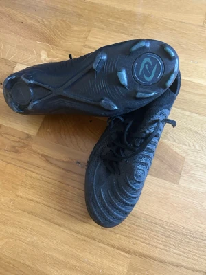 Svarta Nike fotbollsskor med dobbar - Säljer ett par helsvarta Nike phantom elite skor med klassisk snörning och rundad tå. Skorna har mönstrad ovandel i syntetmaterial och fasta dobbar för spel på gräs. Snygg, stilren design med diskret Nike-logga på sidan. Perfekta för dig som vill ha bra grepp och kontroll på planen.