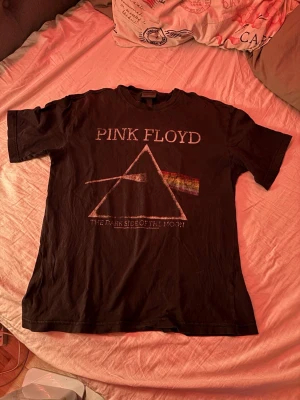 Svart Pink Floyd t-shirt - Klassisk svart t-shirt med Pink Floyds ikoniska 'The Dark Side of the Moon'-tryck på framsidan. T-shirten har korta ärmar och rund hals. Perfekt för dig som gillar bandmerch och vill ha en stilren men ändå uttrycksfull look.