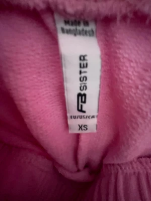 Rosa shorts från FB Sister XS - Säljer ett par ljusrosa shorts från FB Sister i storlek XS. De har elastisk midja och vita kanter längs sidorna för en sportig vibe. Perfekta för sommardagar eller chill hemma. Materialet känns mjukt och bekvämt.