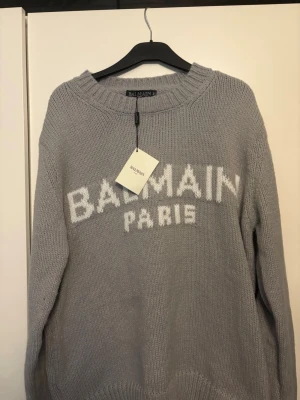 Grå stickad tröja från Balmain - Snygg grå stickad tröja från Balmain med vit logotyptext 'BALMAIN PARIS' framtill. Tröjan har rund halsringning och ribbstickade muddar vid hals, ärmslut och nederkant. Perfekt för dig som gillar exklusiva och trendiga plagg.