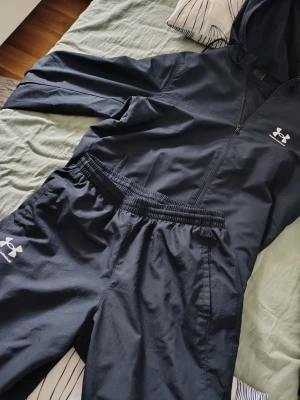 Svart träningsset från Under Armour - Säljer ett svart träningsset från Under Armour med matchande jacka och byxor. Jackan har huva, dragkedja och logga på bröstet. Byxorna har resår i midjan och logga på benet. Perfekt för löpning eller gym. Materialet är lätt och snabbtorkande.