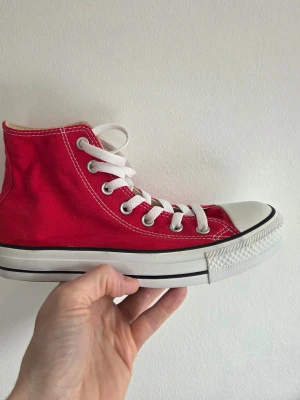 Röda Converse Chuck Taylor Highs - Klassiska röda Converse Chuck Taylor All Star med högt skaft, vita snören och tåhätta i gummi. Skon har vit sula med svart rand och ikonisk logga på sidan. Tillverkad i slitstark canvas och passar perfekt till en streetwear-look.