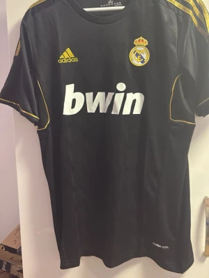 Real Madrid svart matchtröja Adidas - Svart Real Madrid matchtröja från Adidas med gula detaljer och klubbmärke på bröstet. Stort vitt Bwin-tryck på framsidan och ClimaCool-material som andas. Perfekt för fotbollsträning eller att bära som supporter.