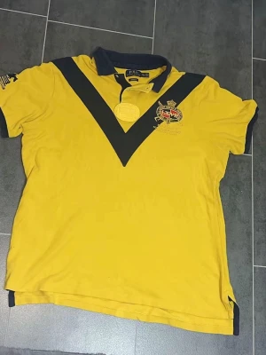 Gul Ralph Lauren t-shirt med tryck - Gul t-shirt från Ralph Lauren med mörkblå detaljer, stor siffra 2 på ryggen och broderad logga på bröstet. T-shirten har korta ärmar, krage och knappslå samt dekorativa knappar på sidorna. Perfekt för dig som gillar sportig stil. Den har en liten defekt, se 3 bild.