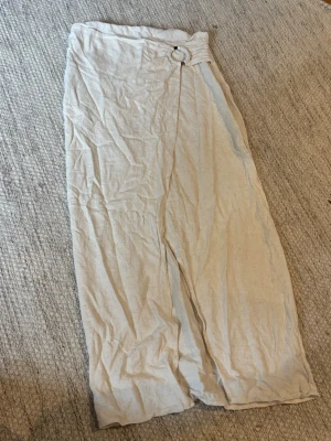 Beige omlottkjol med ringdetalj - Säljer en lång beige omlottkjol i linne med snygg ringdetalj i midjan. Kjolen har en avslappnad passform och är perfekt för varma dagar. Enkel och stilren design som är lätt att matcha med olika toppar. Storlek sitter som S.