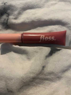 Floss rosa läppglans i tub - Säljer ett snyggt läppglans från Floss i en praktisk tub. Färgen är en mjuk rosa med en hint av nude, perfekt för att ge läpparna en fräsch och glansig look. Tuben är smidig att ha med i väskan och har en enkel applikator. Materialet är plast. Använd fåtal gånger.
