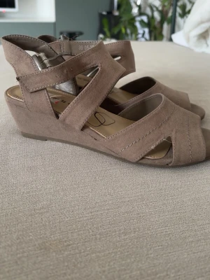 Beige kilklackade sandaler - Snygga beige sandaler med kilklack och öppen tå. Skorna har breda korslagda remmar i mockaimitation och justerbart spänne vid ankeln. Perfekta för en trendig och bekväm look under varma dagar. ENDAST ANVÄND 2 GÅNGER, INGA DEFEKTER ELLER SKADOR