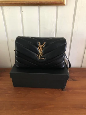 Svart YSL handväska i läder - Snygg svart handväska från Saint Laurent i quiltat läder med guldigt YSL-logo framtill. Väskan har en axelrem och klassisk rektangulär form. Perfekt för dig som vill ha en lyxig accessoar med ikonisk design.