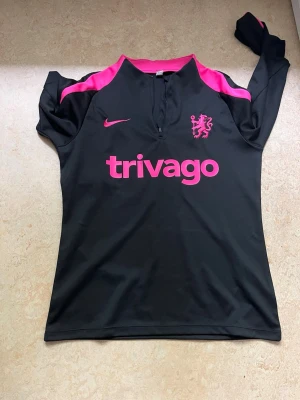 Chelsea träningsjacka svart/rosa Nike - Chelsea FC träningsjacka från Nike i svart med rosa detaljer. Jackan har lång ärm, halv dragkedja och tryck med trivago-logga samt Chelsea-lejonet på bröstet. Tillverkad i ett lätt och stretchigt funktionsmaterial som passar perfekt till träning.