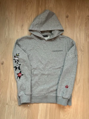 Grå hoodie från Chrome Hearts - Grå hoodie från Chrome Hearts med tryck på bröstet och stora grafiska stjärn- och textmotiv på ärmen och ryggen. Hoodien har huva och ribbade muddar. Materialet är mjukt och bekvämt, perfekt för en avslappnad streetwear-look.
