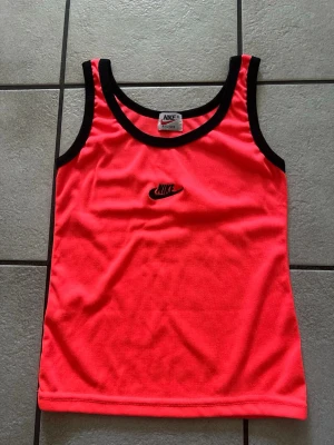 Neonrosa linnetopp från Nike storlek S/M - Snygg neonrosa linnetopp från Nike med svarta kontrastkanter och broderad Nike-logga på bröstet. Perfekt för dig som gillar sportig stil och vill sticka ut. Tillverkad i ett lätt och bekvämt syntetmaterial.
