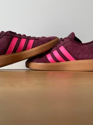 Vinröda Adidas sneakers med rosa ränder - Snygga sneakers från Adidas i vinrött mocka med klassiska rosa ränder på sidorna och brun gummisula. Skorna har lågt skaft och snörning framtill. Perfekta för dig som vill sticka ut med färg och stil. Andvända fåtal gånger. Pris kan diskuteras.
