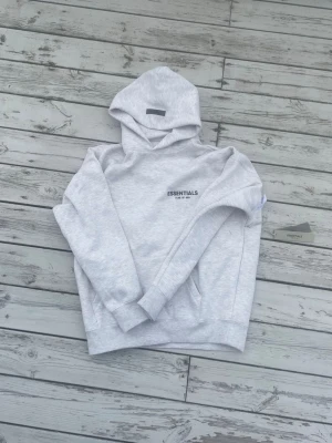 Grå Essentials Fear of God hoodie - Snygg ljusgrå hoodie från Essentials Fear of God med svart logga tryckt på bröstet och ryggen. Hoodien har en klassisk huva, ribbade muddar och känguruficka framtill. Tillverkad i mjukt bomullsmaterial som känns skönt mot huden. Perfekt för en avslappnad streetwear-look.