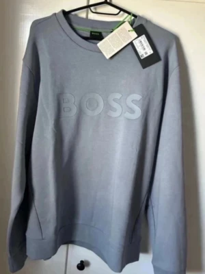 Grå sweatshirt från BOSS - Snygg grå sweatshirt från BOSS med stort ton-i-ton BOSS-tryck på bröstet. Tröjan har rund halsringning, ribbade muddar och är tillverkad i mjukt material som känns skönt mot huden. Perfekt för en avslappnad och trendig look. helt ny