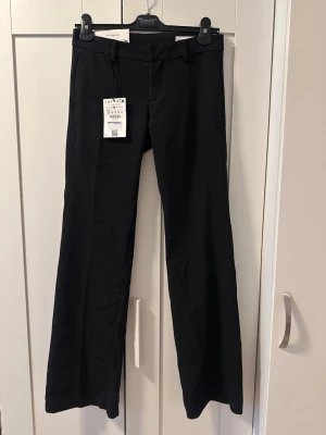 Svarta kostymbyxor - Bershka - Snygga svarta kostymbyxor från Bershka med låg midja. Byxorna har hällor för skärp och klassisk dragkedja med knapp framtill. Perfekta för en clean och stilren look. Materialet är mjukt och följsamt för extra komfort.
