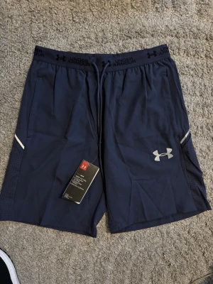 Under armour shorts - Snygga marinblå träningsshorts från Under Armour med elastisk midja och dragsko. Shortsen har sidofickor, meshpaneler för extra ventilation och en vit logga på vänster ben. Perfekta för gymmet eller löpning.