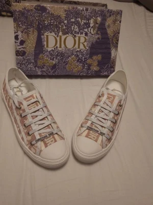 Dior Walk n rosa  - Säljer ett par snygga Dior sneakers i vitt och beige med klassiskt monogrammönster och vita snören. Skorna har en låg siluett, gummisula med stjärndetalj undertill och texten 'Christian Dior' på sidan. Perfekta för dig som vill ha något exklusivt och trendigt.