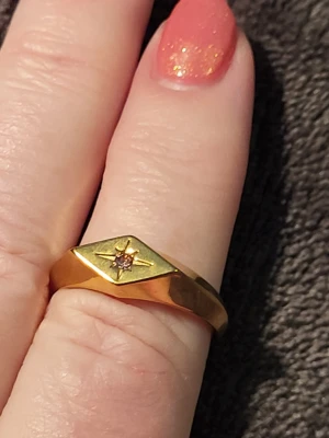 Guldfärgad klackring med stjärndesign - Snygg guldfärgad klackring med en liten ljusrosa sten i mitten av en ingraverad stjärndesign. Ringen har en bredare, kantig ovandel och smalare band runt fingret. Perfekt för dig som gillar unika detaljer och klassisk stil.
