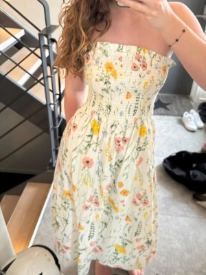 Blommig axelbandslös sommarklänning - Säljer en söt axelbandslös klänning med smock upptill och blommigt mönster i rosa, gul och grön på vit botten. Materialet är bomull och längden är vadlång.