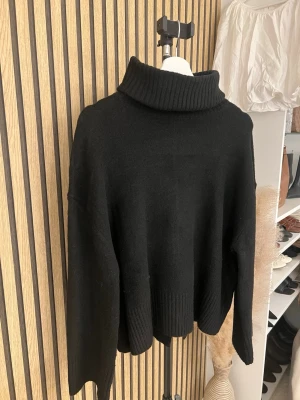 Svart stickad polotröja från pull&bear  - Mysig svart stickad polotröja med bred ribbad krage och avslappnad passform. Tröjan har långa ärmar och ribbade muddar vid ärmslut och nederkant. Perfekt för kyliga dagar och enkel att styla till olika outfits. Använd en gång.