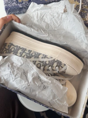 Gråvita Dior sneakers med monogram - Säljer ett par gråvita sneakers från Christian Dior med ikoniskt monogrammönster på sidan. Skorna har vita snören, vit sula och detaljer i canvas och gummi. Klassisk låg modell som ger en clean och lyxig vibe till outfiten.