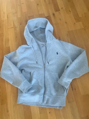 Grå hoodie med dragkedja från Polo Ralph Lauren - Snygg ljusgrå hoodie från Polo Ralph Lauren med dragkedja framtill, huva med dragsko och två fickor. Klassisk broderad logga på bröstet. Perfekt för en avslappnad och stilren look. Storlek S och 170, nypris 2000 kr. 