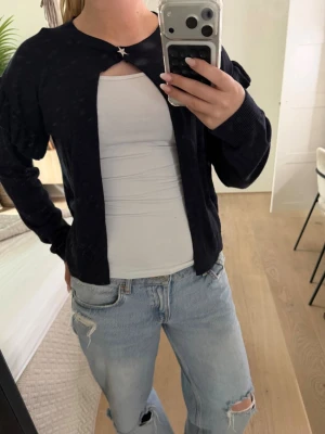 Svart cardigan med volangdetaljer - Svart tunnstickad cardigan med volangdetaljer på ärmarna och en söt stjärnknapp vid halsen. Långärmad och rak passform, perfekt att slänga över ett linne eller t-shirt för en chill look.