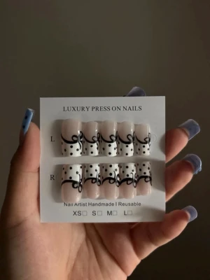 Svartprickiga press-on naglar - Handgjorda press-on naglar i nude med vit bas, dekorerade med svarta prickar och en svart swirl-design. Naglarna har en rak, något fyrkantig form och är återanvändbara. Perfekt för dig som vill ha en unik och trendig look.