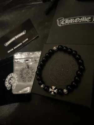 Chrome Hearts svart pärlarmband - STYCK PRIS Snyggt svart pärlarmband från Chrome Hearts med silverdetaljer och ikoniskt korshänge. Armbandet har runda svarta pärlor och flera små silverpärlor med ingraverade mönster. Perfekt accessoar för dig som gillar en edgy och exklusiv stil.