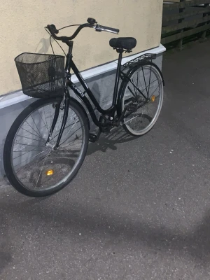 Svart Crescent damcykel med korg - Säljer en svart Crescent damcykel med klassisk ram, stor svart metallkorg fram och pakethållare bak. Cykeln har reflexer på hjulen, bekvämt sadel och raka styren. Perfekt för att ta sig runt i stan eller till skolan.