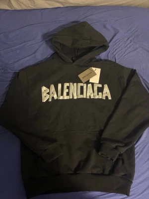 Svart Balenciaga hoodie med tryck - Cool svart hoodie från Balenciaga med stort vitt logotryck på både fram- och baksidan. Klassisk passform med huva och ribbade muddar. Materialet är mjukt och bekvämt, perfekt för en avslappnad streetstyle-look.
