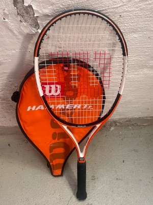 Wilson Hammer Junior tennisracket med fodral - Säljer ett Wilson Hammer Junior tennisracket i orange och vitt med svart handtag och röda strängar. Racketen är av modellen Midplus och kommer med ett matchande orange fodral med stor Wilson-logga.   Mått: Se tredje bilden (66 cm lång, 245 gram). 