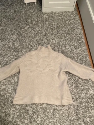 Beige stickad tröja med mönster - Snygg beige stickad tröja med hög krage och diskret rutmönster framtill. Tröjan har långa ärmar och breda ribbade muddar vid ärmslut och nederkant. Perfekt för lager-på-lager och en stilren look. Är i storlek M men är liten i storlek. 