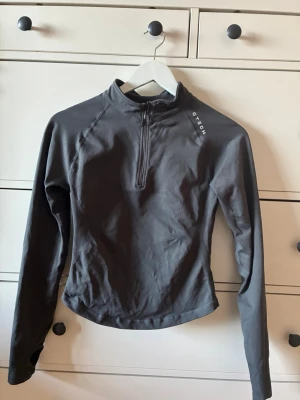 Mörkgrå långärmad träningströja GTECH - Mörkgrå långärmad träningströja från GTECH med halv dragkedja framtill och hög krage. Tillverkad i ett stretchigt funktionsmaterial som andas. Tröjan har tumhål vid ärmsluten och en smal, kroppsnära passform. Perfekt för löpning eller gym.