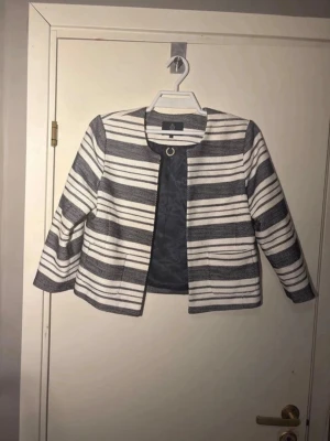 Randig kavaj från Stockh LM - Snygg kavaj från Stockh LM i vitt och mörkblått med breda horisontella ränder. Kavajen har rundad halsringning, trekvartsärmar och ett dekorativt guldfärgat spänne framtill. Fodrad insida för extra komfort.