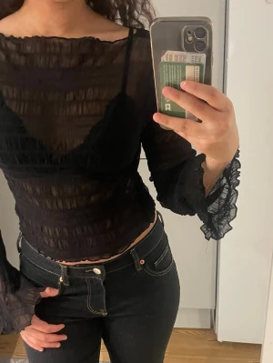 Svart mesh blus med volangärm - Trendig svart mesh blus med transparent material och volangdetaljer vid ärmslut. Blusen har lång ärm och croppad passform, perfekt för att skapa en edgy look. Snygg att styla med både jeans och kjol.