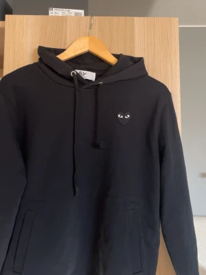 Svart hoodie från Comme des Garçons - Svart hoodie med huva från Comme des Garçons Play. Ikoniskt hjärtlogo med ögon broderat på bröstet. Mjuk bomullskänsla och fickor på framsidan. Perfekt för dig som gillar stilrena och trendiga plagg.