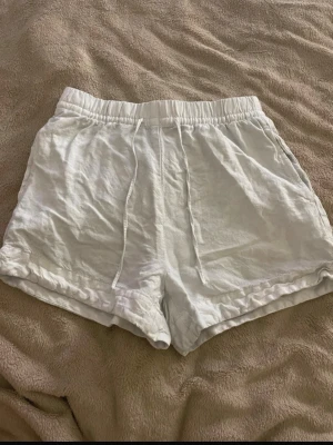 Vita linne shorts med snörning - Säljer ett par vita shorts i linne med elastisk midja och snörning. De har en loose passform och uppvikta benslut, perfekta för varma dagar. Shortsens material är lätt och luftigt, vilket gör dem riktigt sköna att bära.