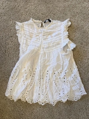 Vit broderad blus med volangärm - Söt vit blus med broderade detaljer och hålmönster. Blusen har volangärmar, rund halsringning och knäppning bak i nacken. Tillverkad i ett lätt bomullstyg och vågig kant nedtill. Blusen har gått ur sortimen.